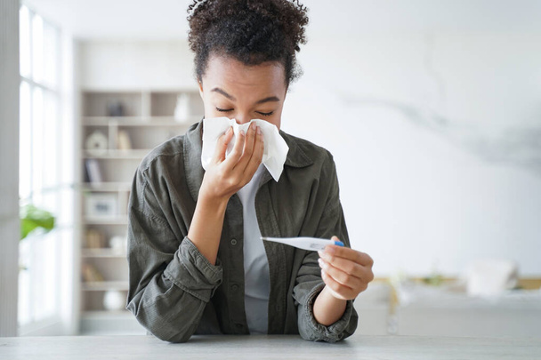 Teenage afro girl caught cold. Young hispanic woman is measuring temperature with thermometer, sneezing and blowing running nose. Flu or coronavirus symptoms. Girl is staying home while sick. - Photo, image