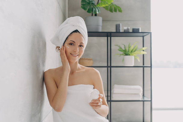 Carefree young woman is relaxing in bathroom. European lady applies face cream. Attractive caucasian girl wrapped in towel after bathing and hair washing. Happy girl at spa resort. - 写真・画像