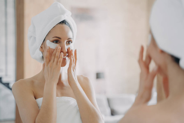 Girl applies eye patches and looking into mirror. Attractive european woman wrapped in towel after bathing. Young hispanic lady takes shower at home. Relaxation at spa resort. Beauty routine at home. - Φωτογραφία, εικόνα