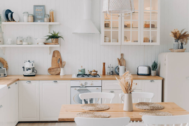 Scandinavian kitchen design. Modern apartment decoration. Domestic kitchen or dining room. White luxurious interior lit by daylight. Kitchen appliance, utensils, stove, table and refrigerator. - Photo, Image