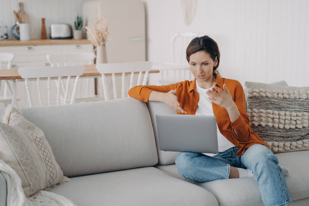 Young hispanic woman has online conference at home. Girl with laptop is talking at internet meeting. Pretty lady is sitting on sofa having discussion. Working while quarantine. - Foto, Imagen
