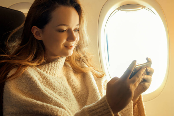 A beautiful girl is sitting in an airplane looking at the phone, reading a book or playing games. Air travel, long flight. - Photo, Image