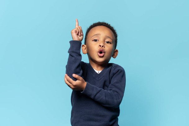 Niño negro divertido niño sosteniendo el dedo hacia arriba y mirando a la cámara, mostrando una nueva idea en el fondo del estudio azul. Adorable niño inteligente y espacio de copia para su texto - Foto, imagen