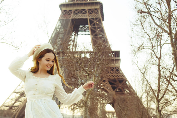 Gyönyörű szőke Párizsban az Eiffel-toronnyal a háttérben svájcisapkában és fehér ruhában. A nő nevet és mosolyog, a lány Franciaországba utazik.. - Fotó, kép