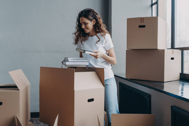 Curly young woman, student is housing in residential room. Happy european woman unpacking boxes with books. Girl in white t-shirt indoors. Relocation and independence concept. - Foto, Imagem