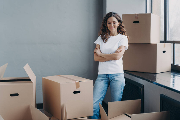 Satisfied woman unpacking boxes. Attractive girl in new apartment. Young woman in jeans and white t-shirt. Relocation, delivery service and new apartment concept. - Photo, Image