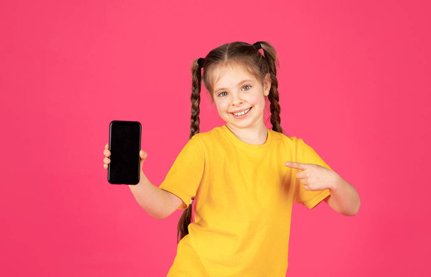 Sorridente bambina puntando Smartphone vuoto con schermo nero in mano, carino allegro Preteen Femmina bambino raccomandare una nuova applicazione mobile mentre in piedi su sfondo rosa, Mockup - Foto, immagini