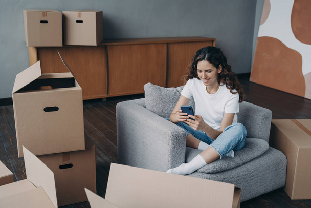 Young spanish woman posting news. Happy apartment buyer unpacking boxes. Girl is moving to new home. Relaxed young lady clicks mobile phone. Independence and achievement concept. - Fotoğraf, Görsel