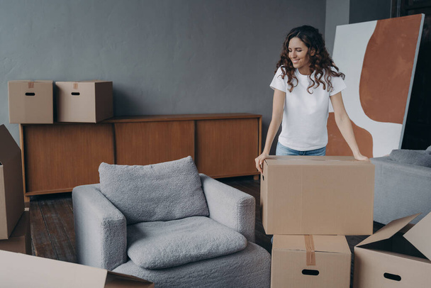 Smiling european woman is packing things. Girl moves to new house. Young lady is unpacking cardboard boxes in new apartment. Big new living room with armchair. Real estate purchase concept. - Foto, immagini
