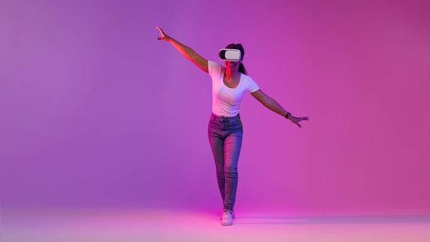 Веселая черная женщина в наушниках виртуальной реальности исследуя деятельность в киберпространстве, прогуливаясь в неоновом свете над фиолетовой студией, взволнованная афроамериканская женщина балансирует, делая шаги, копируя пространство - Фото, изображение