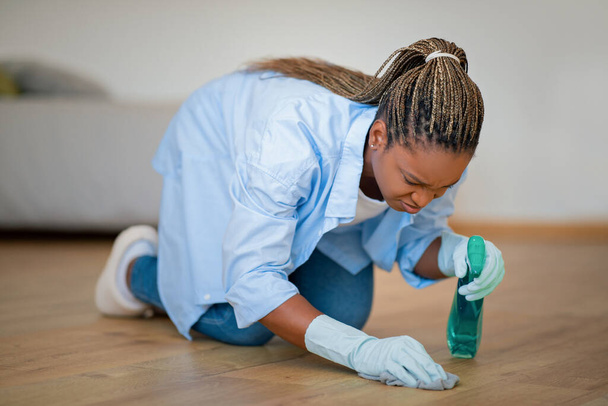 Annoyed young black woman housewife or maid standing on knees, erasing removing dirt stain from wooden floor, using cleaning spray and duster cloth, home imterior, copy space - Φωτογραφία, εικόνα