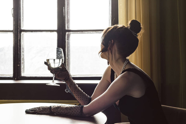 Piękna, piękna i elegancka dziewczyna w czarnej sukience i rękawiczki Mesh Sheer na rękach, siedząca przy stole ze szklanką w dłoni. Pojęcie elegancji i wyrafinowania. Impreza formalna lub ekskluzywna. - Zdjęcie, obraz