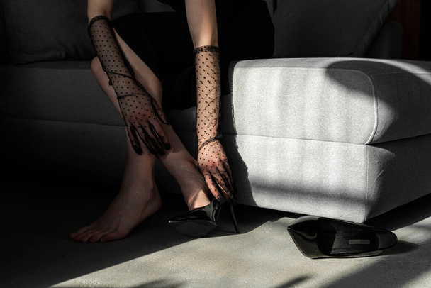 Een elegant meisje in een zwarte jurk en Mesh Sheer handschoenen aan haar handen, trekt haar schoenen uit van vermoeide voeten na een lange dag. Concept van elegantie en verfijning. Formele of belangrijke gebeurtenis. - Foto, afbeelding