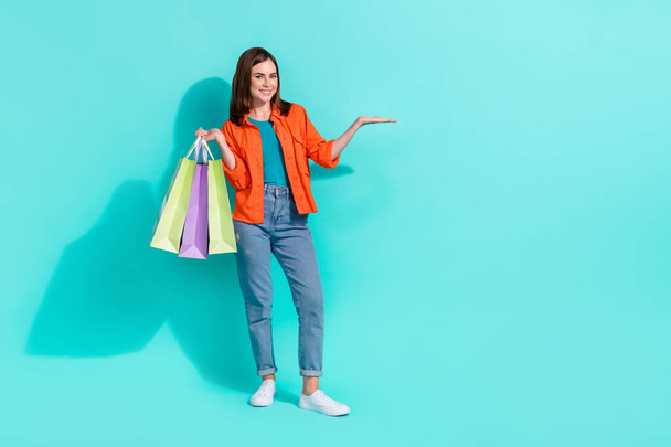 Full length photo of cheerful cute girl dressed shirt rising shoppers holding arm empty space isolated turquoise color background. - Foto, Imagem