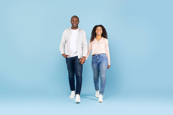 Afrikai amerikai pár fogja egymás kezét, és séta felé kamera, középkorú férfi és fiatal nő pózol kék stúdió háttér, teljes test hossza, másolás tér - Fotó, kép