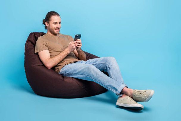 Full length photo of cute cheerful man wear beige t-shirt bean bag texting modern gadget emtpy space isolated blue color background. - Foto, Imagem