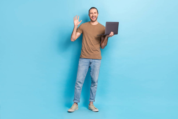 Full length photo of positive friendly man wear beige t-shirt waving arm talking modern gadget isolated blue color background. - Photo, Image