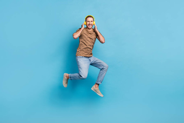 Full length photo of excited cheerful man wear beige t-shirt jumping high enjoying music empty space isolated blue color background. - Φωτογραφία, εικόνα