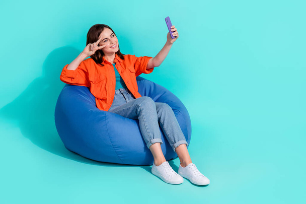 Full length photo of cute pretty girl dressed shirt bean bag showing v-sign device empty space isolated turquoise color background. - 写真・画像