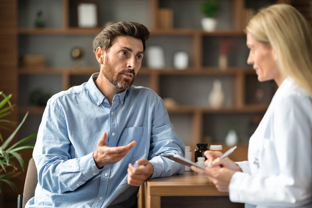 Male Patient Sharing Health Problems With Doctor Woman During Meeting In Office, Upset Sick Man Talking To Therapist Lady, Female Physician Holding Clipboard And Making Notes, Selective Focus - Foto, Imagem
