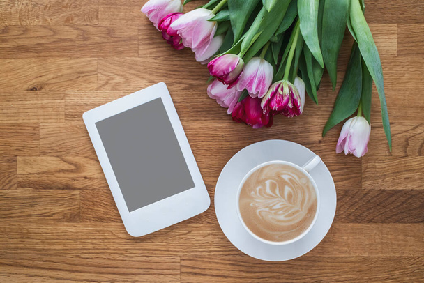Kopje koffie met roze tulpen en e-reader op houten ondergrond. - Foto, afbeelding