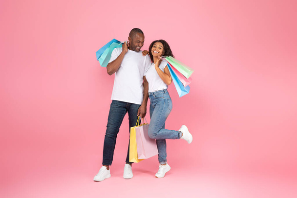 Großer Verkauf und Shopping mit Vergnügen. Aufgeregtes schwarzes Paar hält viele farbige Pakete in Händen, posiert isoliert auf rosa Hintergrund, Studioaufnahme, volle Länge - Foto, Bild