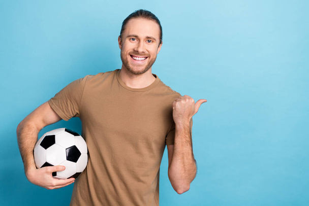 Foto de buen humor bastante hombre usar camiseta beige sosteniendo pelota de fútbol pulgar espacio vacío aislado color azul fondo. - Foto, Imagen