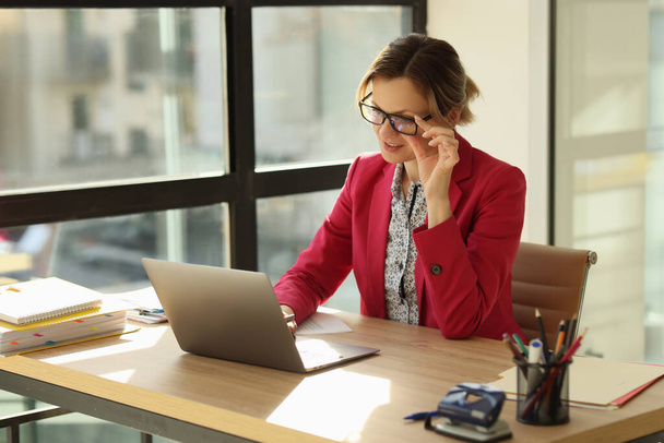Female manager sits at her workplace in office, touches her glasses and looks at laptop screen. Office work and online business concept. - Photo, Image