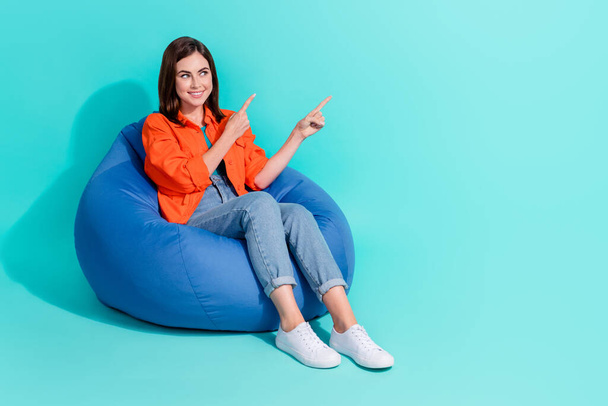 Full length photo of dreamy shiny lady wear orange shirt sitting bean bag pointing empty space isolated teal color background. - Fotó, kép