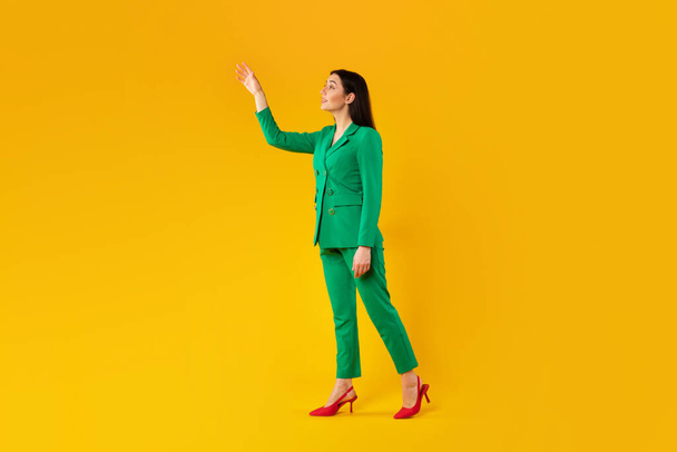 Κομψό νεαρή κοπέλα αγγίζει αόρατο αντικείμενο, δείχνει και διαφημίζει κάτι, στέκεται πάνω από κίτρινο φόντο στούντιο, πλήρες μήκος, ελεύθερο χώρο - Φωτογραφία, εικόνα