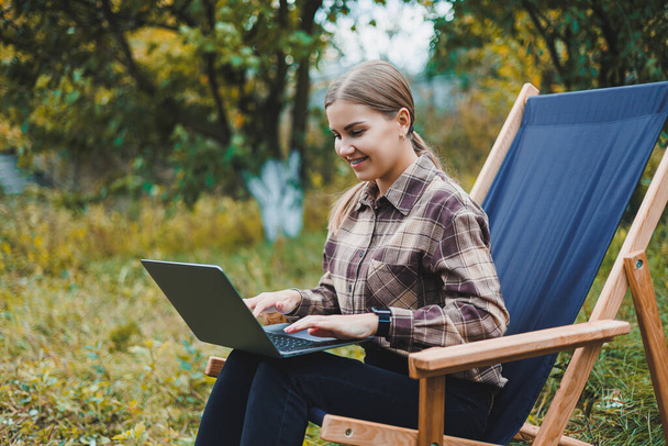 Schöne Frau im karierten Hemd mit Laptop arbeitet draußen im Garten, Homeoffice-Konzept. Fernbedienung. - Foto, Bild