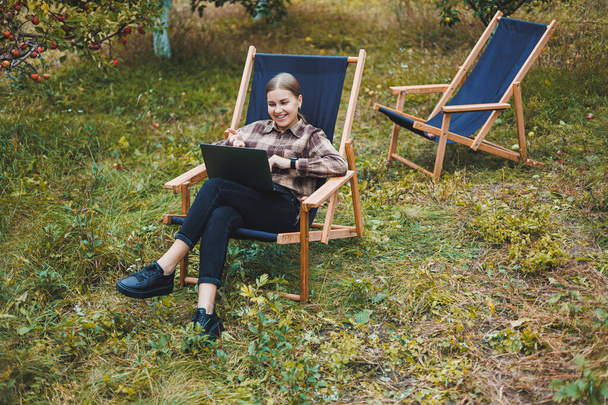 Beautiful woman in checkered shirt with laptop working outdoors in garden, home office concept. Remote work. - Fotó, kép