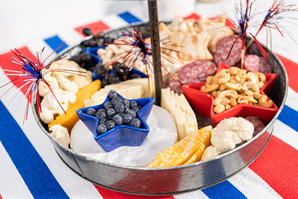 4 de julho placa de charcutaria em um suporte de metal de duas camadas, cheio de queijo, biscoitos, salame e frutas frescas - Foto, Imagem