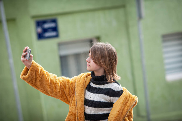 Young pretty woman in a yellow fur coat, social media influencer, walk the streets of the city with her black dog, using smartphone. - Fotoğraf, Görsel
