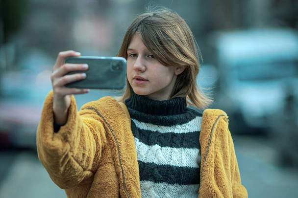 Young pretty woman in a yellow fur coat, social media influencer, walk the streets of the city with her black dog, using smartphone. - Fotoğraf, Görsel