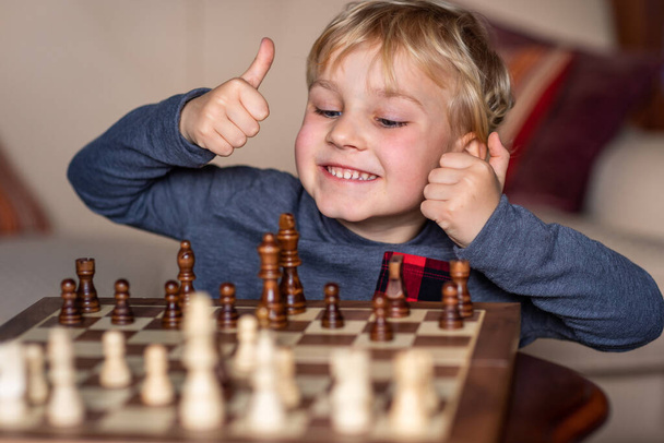 Small child 5 years old playing a game of chess on large chess board. Chess board on table in front of the boy he is happy to win, showing thumbs up - Fotoğraf, Görsel