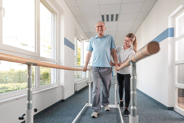 Senior Patient and physical therapist in rehabilitation walking exercises, she is helping him along the bars - Photo, Image