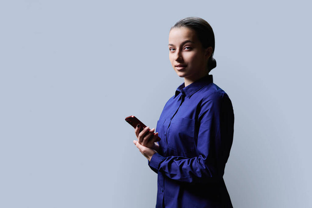 Teenage girl using smartphone, smiling female looking at camera, gray background copy space. High school teen student, goods services educational programs for teenagers - Foto, immagini