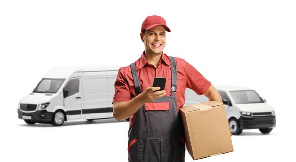Courier in a uniform holding a box and using a mobile phone in front of vans isolated on white background - 写真・画像
