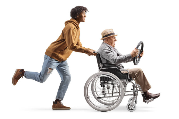 Full length profile shot of a young african american man pushing a mature man in a wheelchair holding a steering wheel isolated on white background - Фото, изображение