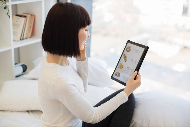 Casually dressed woman holding digital tablet and having mobile conversation while sitting on cozy white bed. Happy lady staying at home and ordering food for lunch in online store with discount. - Fotoğraf, Görsel