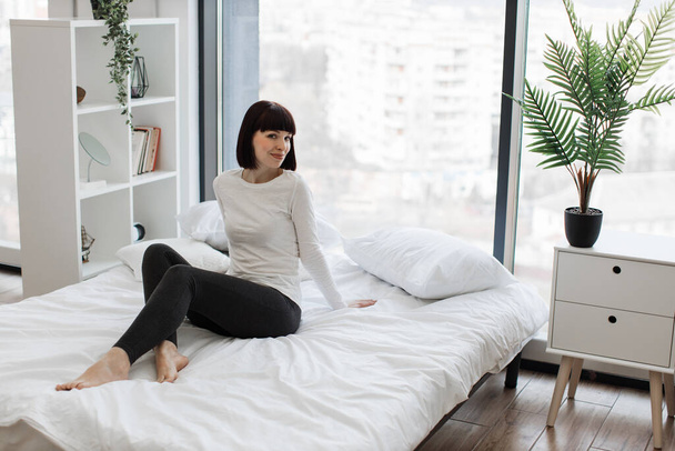 Attractive caucasian woman in domestic outfit resting on white bed with panoramic windows on background. Leisure time at home and lifestyles concept. - Foto, afbeelding