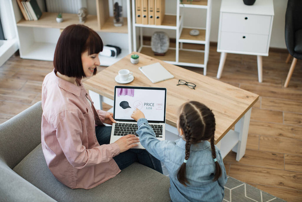 Rear view of dark haired woman sitting on couch with laptop on knees while her pretty girl pointing on screen with forefinger. Mother and daughter choosing new clothes to buy during discounts. - Foto, Imagen