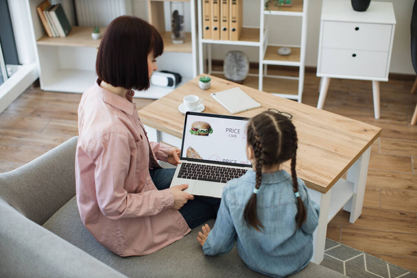 Dark haired woman and little girl using portable laptop for ordering lunch in online shop. Happy mother with lovely daughter purchasing food with discount for saving and free time at home. - Foto, immagini