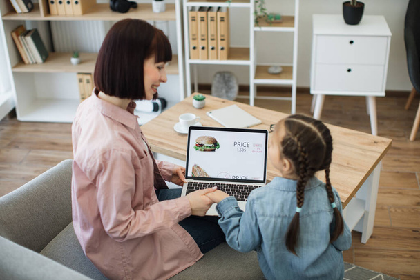 Dark haired woman and little girl using portable laptop for ordering lunch in online shop. Happy mother with lovely daughter purchasing food with discount for saving and free time at home. - Foto, Imagen