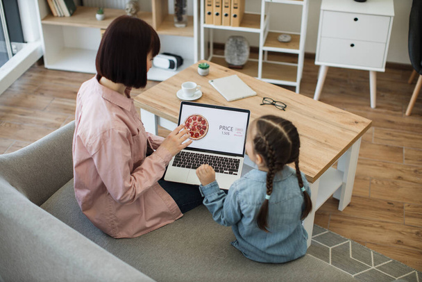 Happy mother and cute little daughter purchasing tasty pizza with discount of 65 percent using wireless laptop for ordering takeaway food. Woman and girl sitting together on couch at cozy living room. - Fotografie, Obrázek