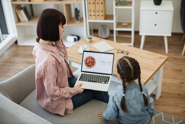 Beautiful caucasian woman holding laptop and talking on smartphone while sitting with pretty daughter on couch. Happy family of two making online order of pizza with discount 65 percent - Foto, afbeelding