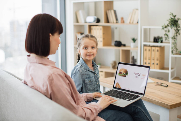 Side view of charming brunette sitting on couch with daughter and using wireless laptop. Cute little girl smiling and looking at camera while focused mother scrolling online shop with fast food. - Φωτογραφία, εικόνα