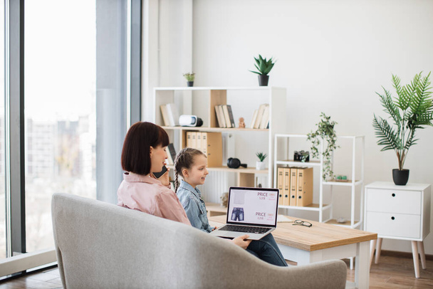 Adorable little girl sitting on couch with her young mother while buying new clothes with discount in internet. Modern caucasian family using smartphone and laptop for online purchase. - Foto, Bild