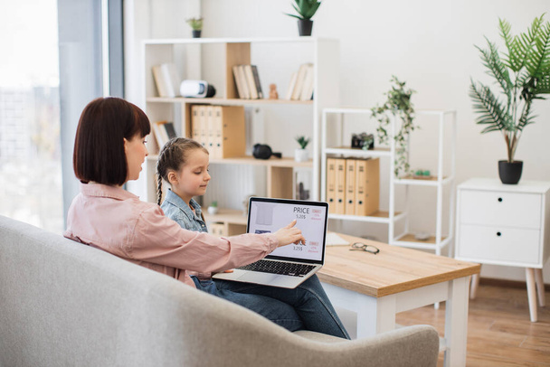 Caring mother with little daughter browsing ecommerce site for searching discount of new trendy apparel. Young woman and pretty girl sitting together on comfy couch and using modern wireless laptop. - Φωτογραφία, εικόνα